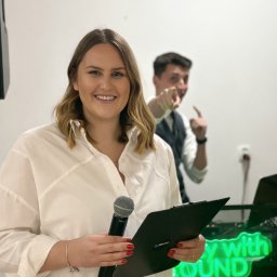 OLSOUND Wiktor Olszewski - Uśmiechnięta prezenterka z mikrofonem i clipboardem na tle neonu 'Play with Sound', za nią mężczyzna wskazujący palcem, sugerujący interakcję z publicznością.