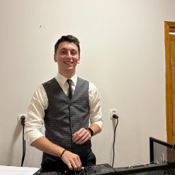 OLSOUND Wiktor Olszewski - Uśmiechnięty DJ w kamizelce obsługuje konsoletę na tle białej ściany z gniazdkami i fragmentem drewnianych drzwi. Widoczny neonowy napis.