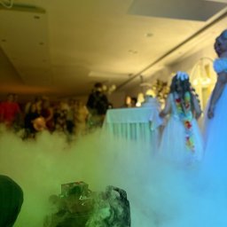 OLSOUND Wiktor Olszewski - Efektowna para z wytwornicy dymu na sali balowej podczas uroczystości, w tle goście i tort weselny. Oświetlenie sceniczne tworzy magiczną atmosferę.