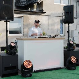 OLSOUND Wiktor Olszewski - DJ w okularach i czapce obsługujący białą konsolę z drewnianym blatem, otoczony profesjonalnym nagłośnieniem Yamaha i ruchomymi głowami oświetleniowymi na zielonym dywanie, na tle domu.