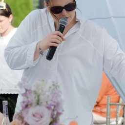 OLSOUND Wiktor Olszewski - Kobieta w białej koszuli i okularach, z mikrofonem Shure w dłoni, występuje na świeżym powietrzu. Na pierwszym planie rozmyte kwiaty i zabawki na stole.