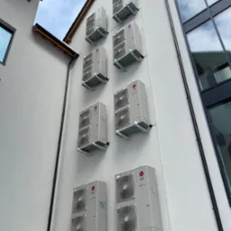 Fasada budynku z zamontowanymi jednostkami zewnętrznymi klimatyzacji LG Multi V, widoczne białe rurki instalacyjne. Ujęcie z dołu, budynek z nowoczesną architekturą.