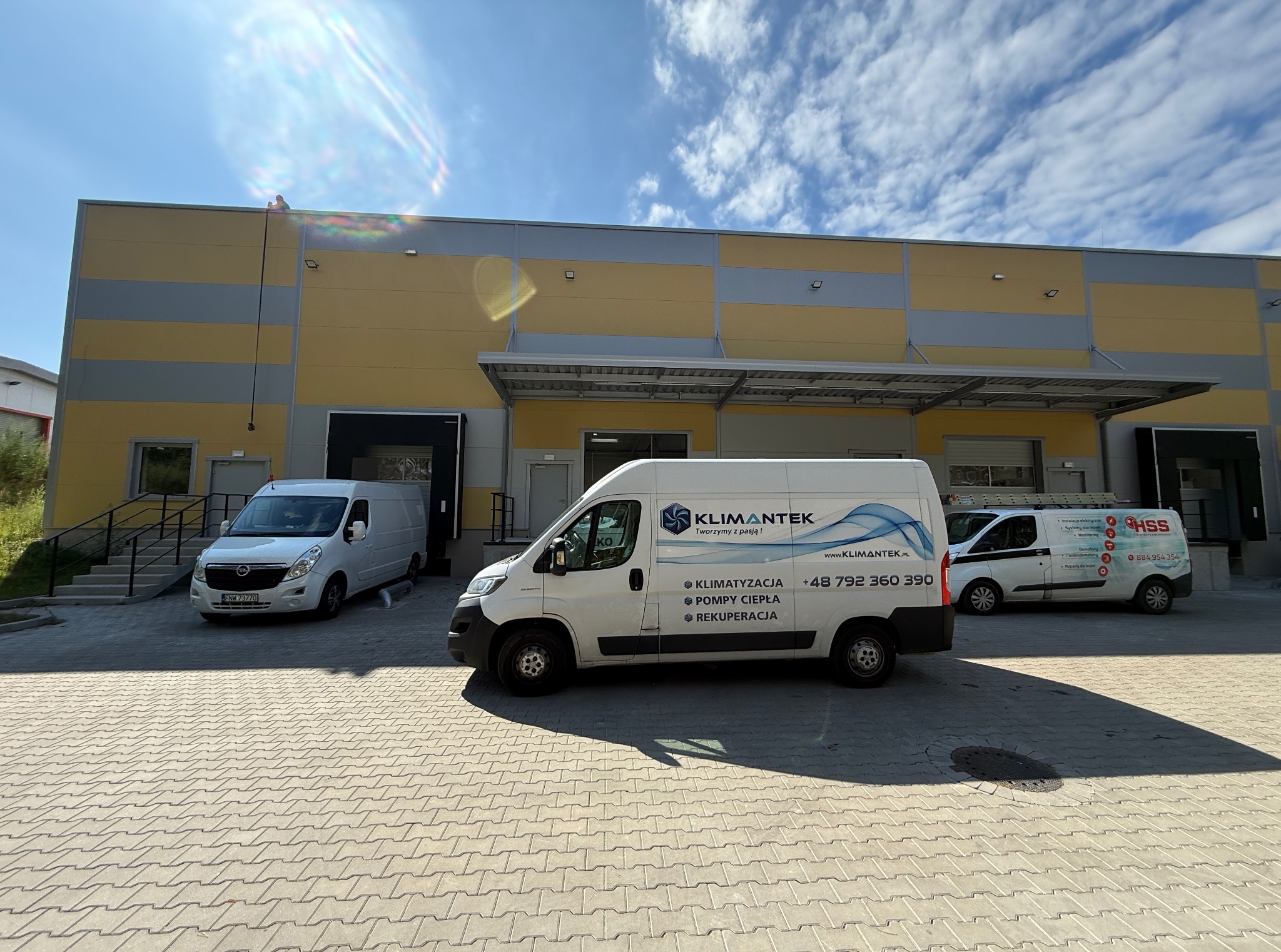 Flota białych samochodów dostawczych z logo Klimantek na tle magazynu. Usługi: klimatyzacja, pompy ciepła, rekuperacja. Błękitne niebo z chmurami w tle.
