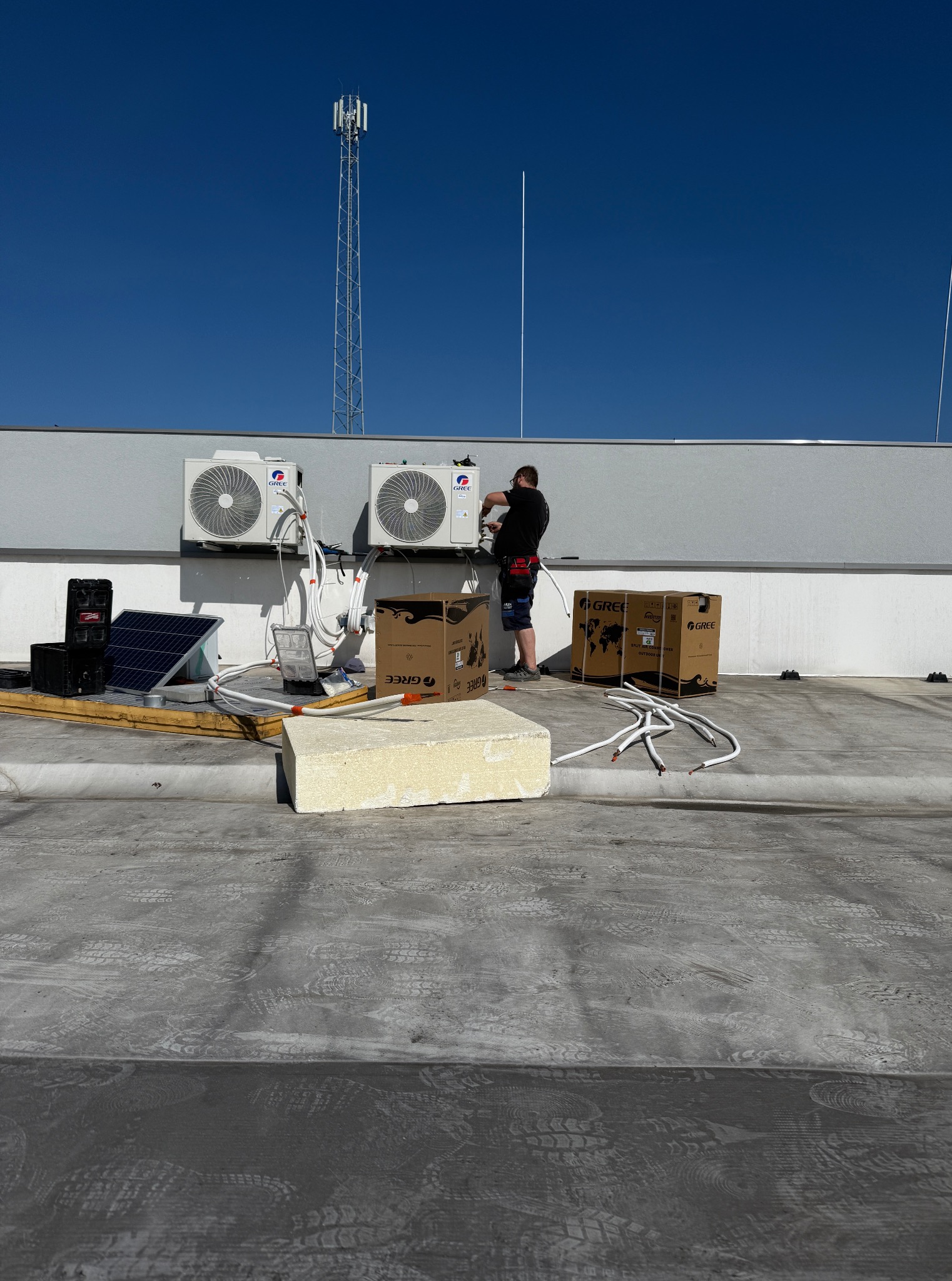 Instalacja klimatyzacji na dachu budynku: monter łączy jednostki zewnętrzne Gree, widoczne skrzynki, przewody i antena telekomunikacyjna w tle.