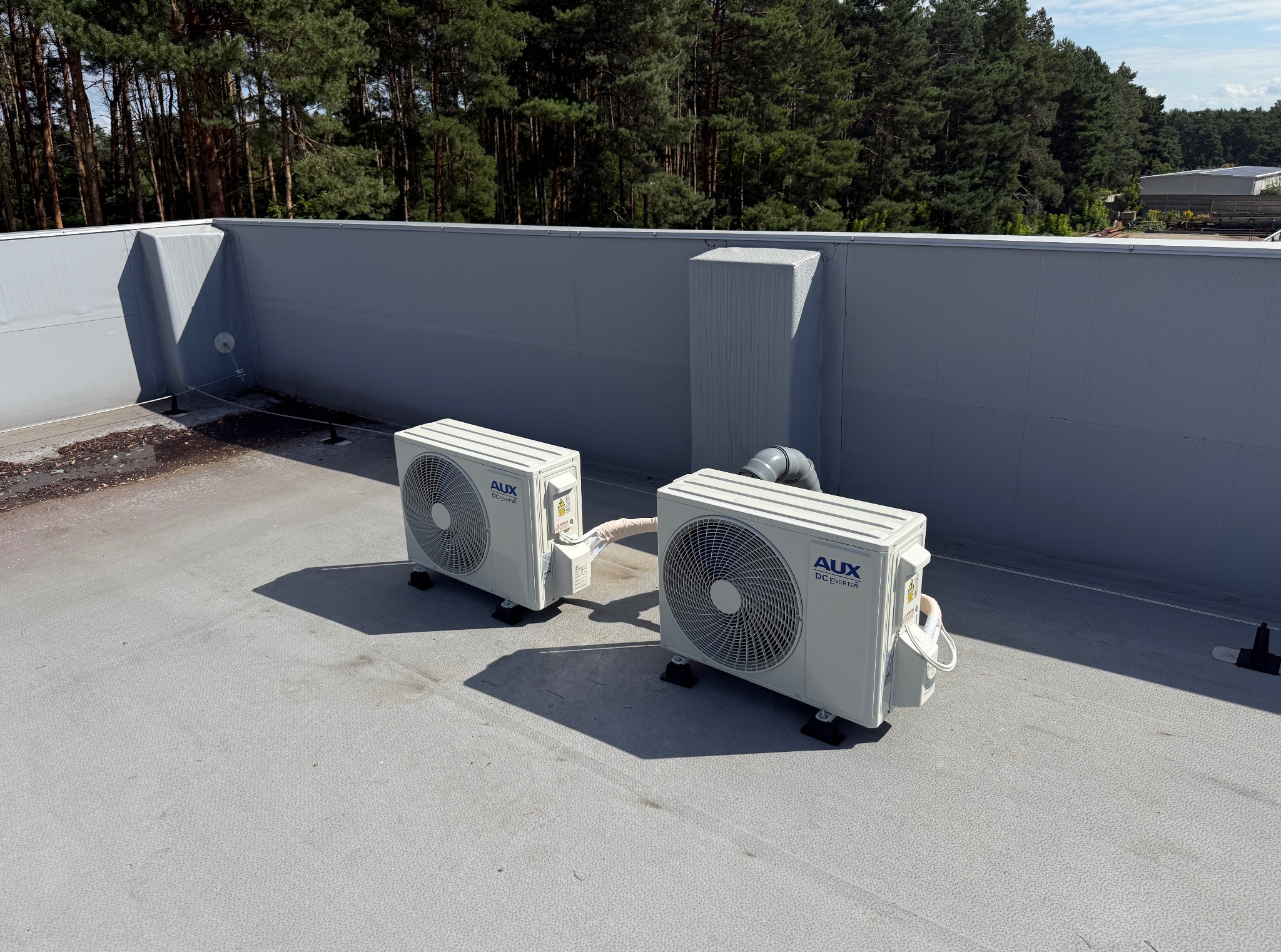 Dwa klimatyzatory AUX DC Inverter na szarym dachu budynku z widokiem na las iglasty w tle. Instalacja z widocznymi przewodami i elementami mocującymi.