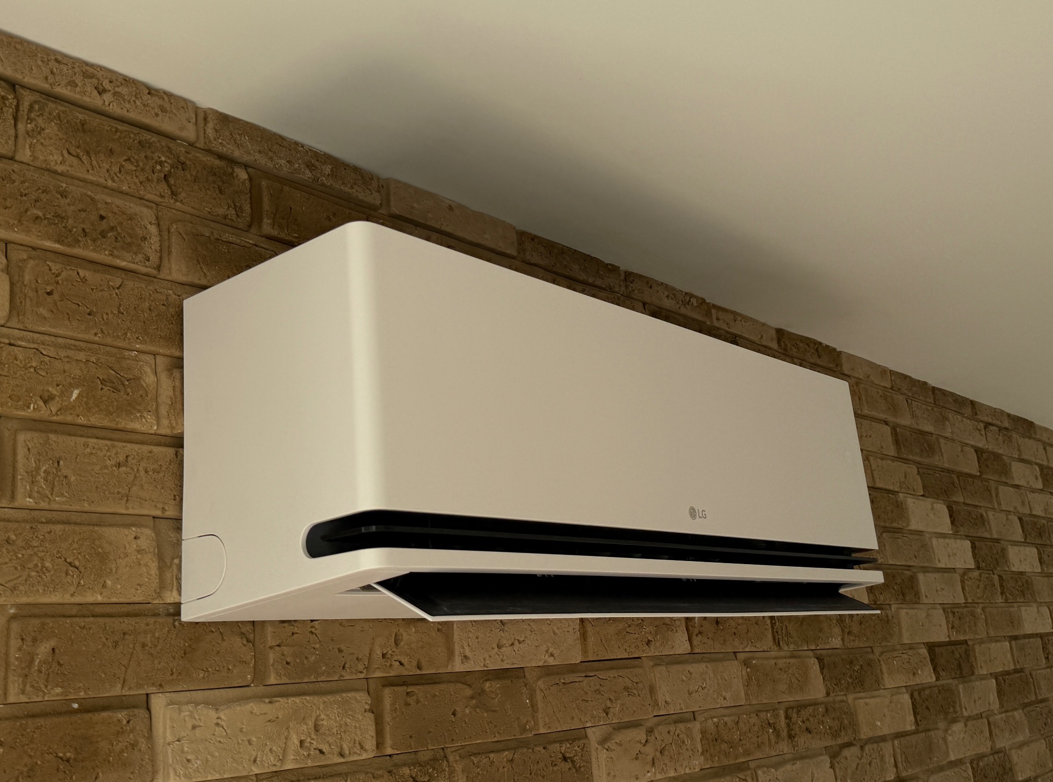 Biała klimatyzacja LG na ścianie z cegły, widok z dołu. Otwarta klapa nawiewu, widoczne logo LG. Oświetlenie naturalne, ciepłe barwy cegły.