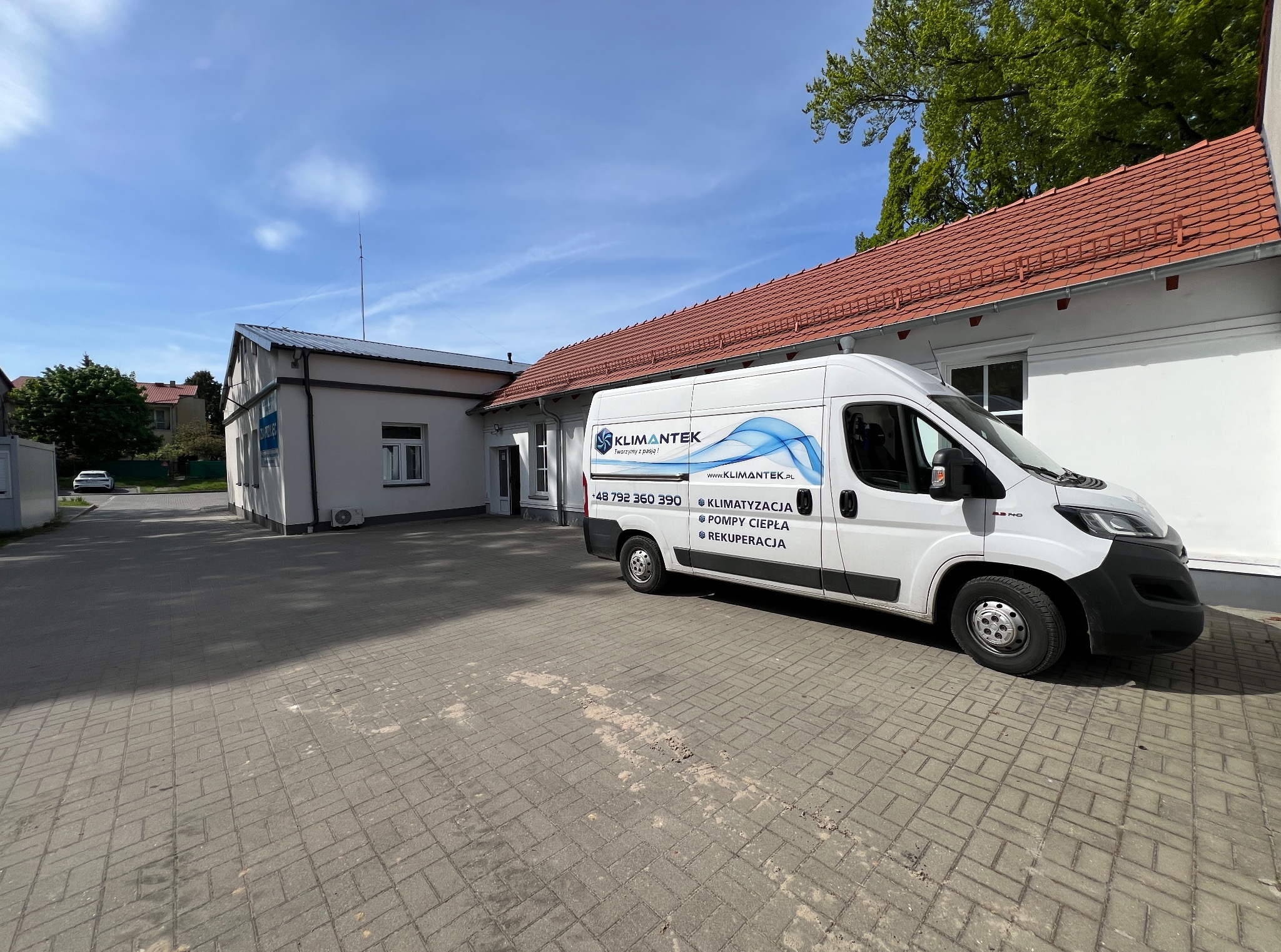 Biały samochód dostawczy firmy Klimantek zaparkowany przed budynkiem z czerwoną dachówką. Na aucie logo i informacje o klimatyzacji, pompach ciepła i rekuperacji.
