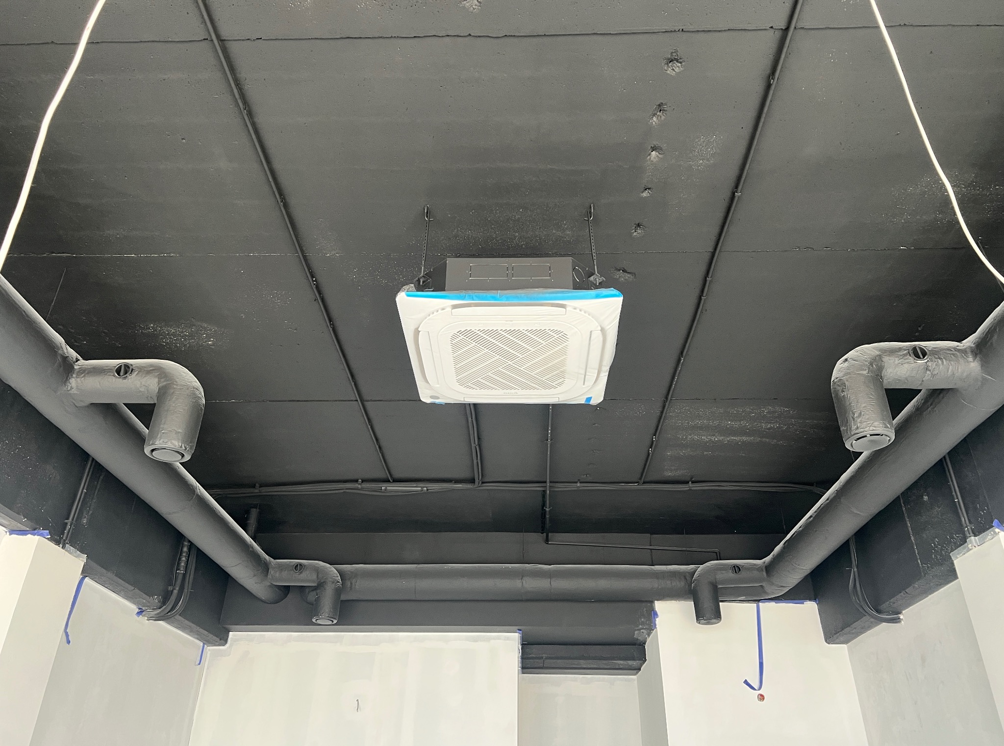 Sufit z instalacją: czarny sufit, biała klimatyzacja kasetonowa z niebieską folią ochronną, szare rury wentylacyjne. Widok z dołu, surowy stan wykończenia.