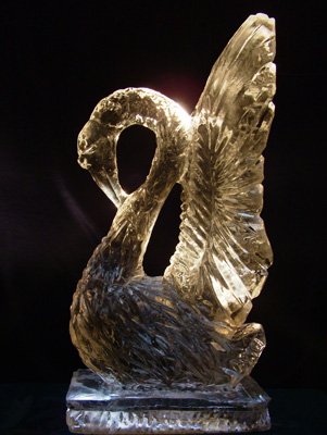 Lodowa rzeźba przedstawiająca łabędzia z uniesionymi skrzydłami, umieszczona na lodowej podstawie, oświetlona od góry, na ciemnym tle.