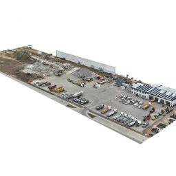 L-PROJEKT USŁUGI PROJEKTOWE I INŻYNIERSKIE Paweł Lipski - Szeroki widok z lotu ptaka na plac budowy z budynkiem magazynowym, ciężarówkami, maszynami budowlanymi i składowanym materiałem. Widoczne panele słoneczne na dachu budynku.