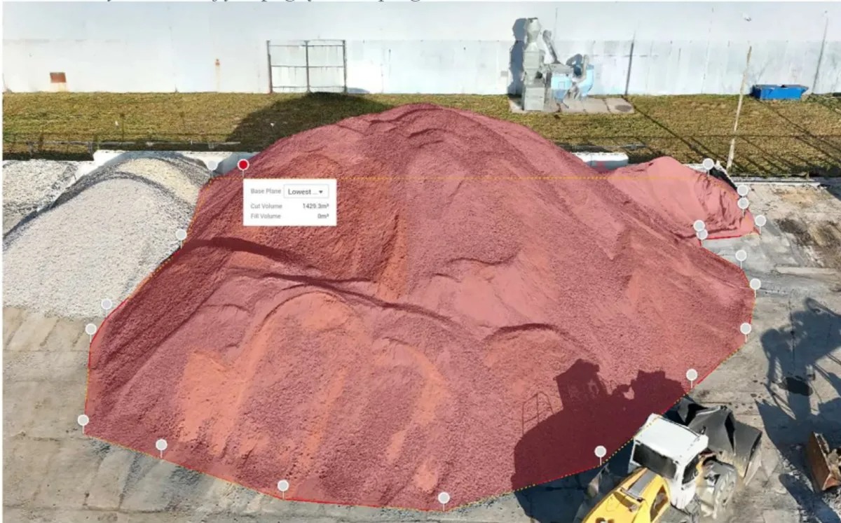 Pomiar objętości hałdy kruszywa z użyciem drona. Nałożona siatka pomiarowa z punktami kontrolnymi. Widoczny sprzęt budowlany i fragment budynku w tle.