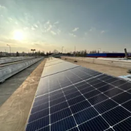 Instalacja paneli słonecznych na płaskim dachu budynku komercyjnego, widok z góry, w tle błękitne niebo i budynki. Słoneczny dzień, równomierne rozmieszczenie paneli.