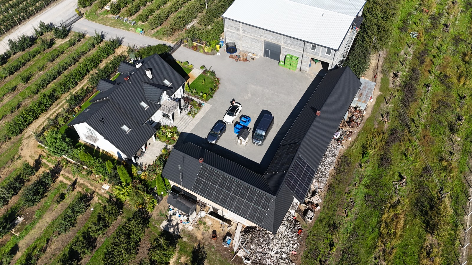 Widok z drona na dom z panelami słonecznymi w Pruszkowie, otoczony sadem, z zaparkowanymi autami i budynkiem gospodarczym. Nowoczesna instalacja OZE.