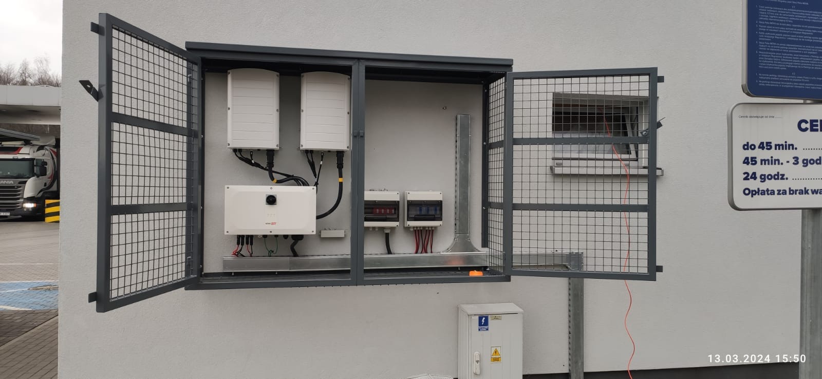 Zewnętrzna instalacja fotowoltaiczna w metalowej szafce z otwartymi drzwiczkami, widoczne falowniki, zabezpieczenia i okablowanie. Instalacja przy ścianie budynku.