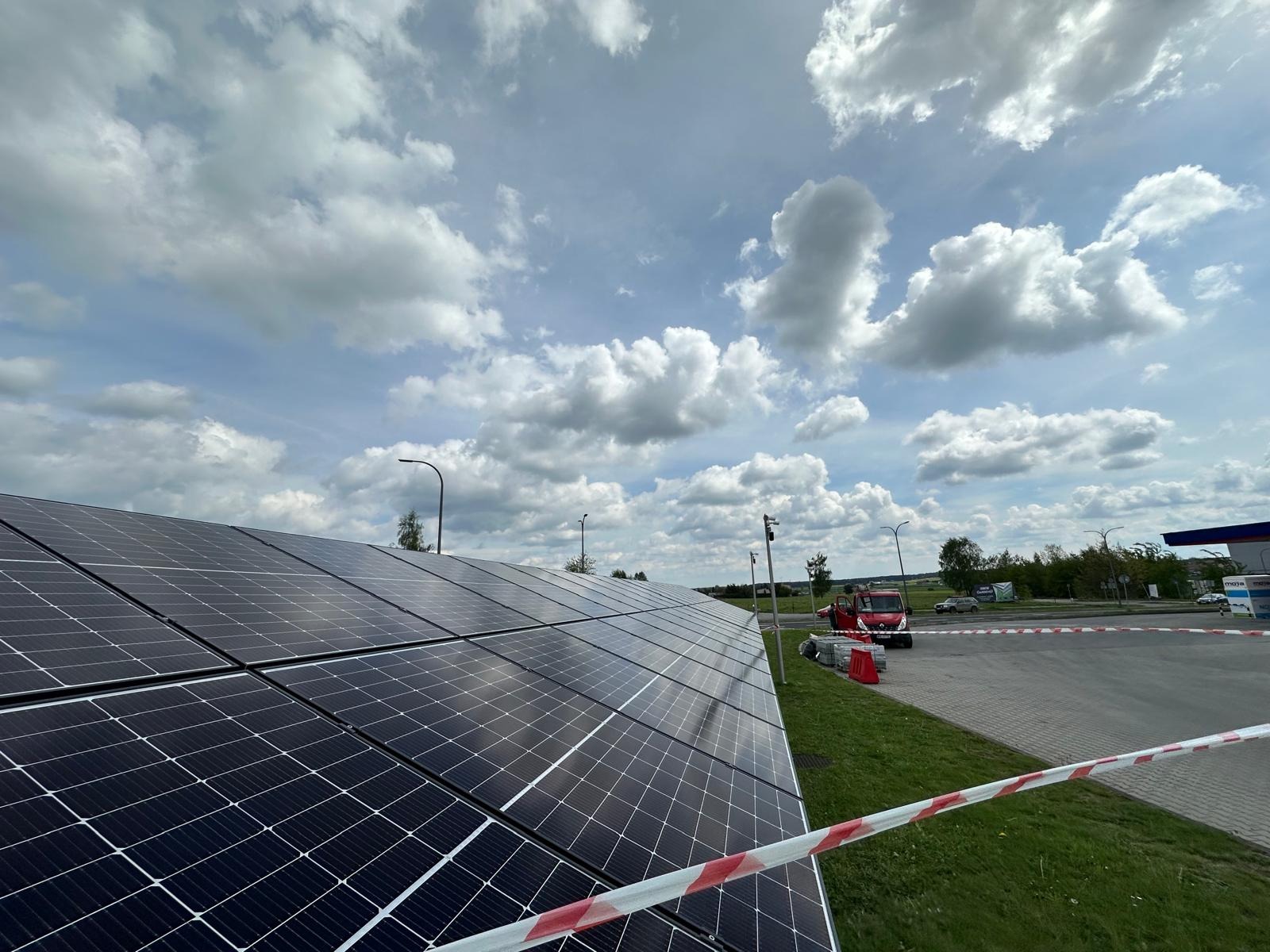 Instalacja paneli słonecznych z bliska, widoczne niebo z chmurami, budynek stacji paliw w tle, taśma ostrzegawcza na pierwszym planie. Ujęcie z dołu.