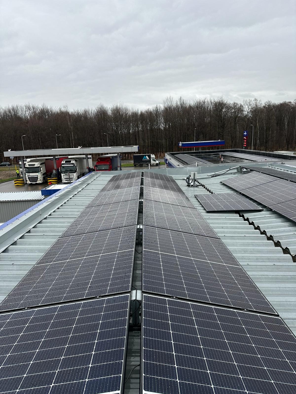 Instalacja fotowoltaiczna na dachu budynku komercyjnego, widok z góry. Panele w kroplach deszczu, w tle stacja benzynowa i las.