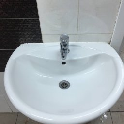 Tidytigers - Biała umywalka z chromowaną baterią na tle płytek. Widoczne zabrudzenia na fugach i podłodze, co sugeruje potrzebę sprzątania.