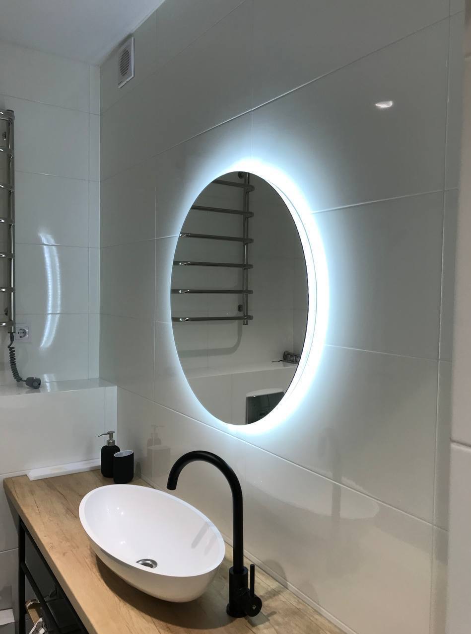 Nowoczesna łazienka: owalne lustro z podświetleniem LED, umywalka nablatowa na drewnianym blacie, czarna bateria i białe płytki na ścianach.