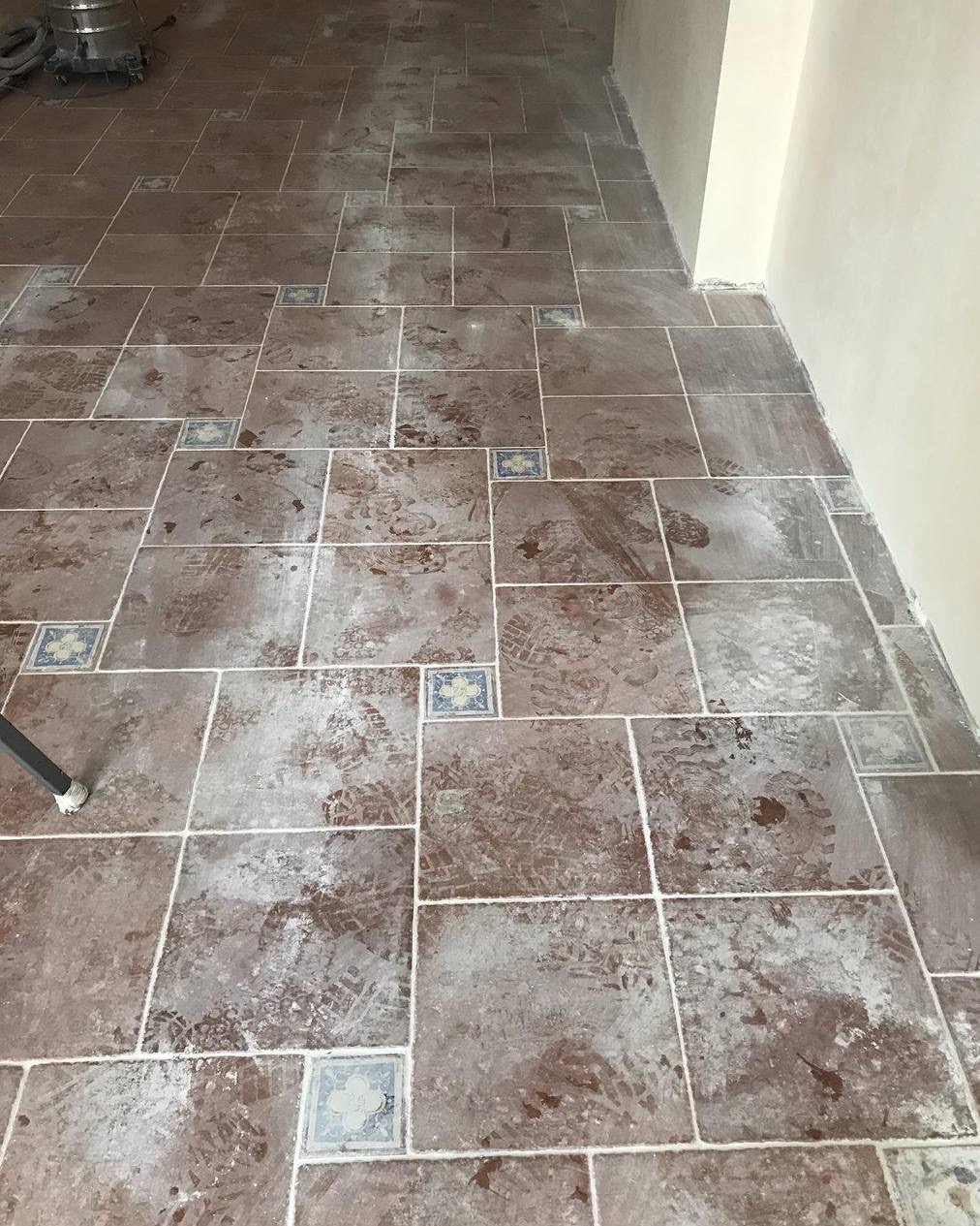 Brudna, brązowa podłoga z płytek z dekoracyjnymi wstawkami, pokryta śladami butów i resztkami po remoncie. Widoczny fragment odkurzacza przemysłowego.