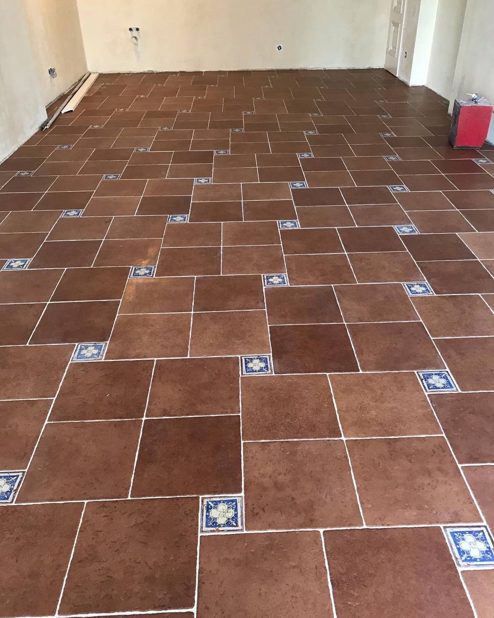 Ułożona podłoga z brązowych płytek z dekoracyjnymi, niebieskimi wstawkami i białymi fugami. Widok na całą powierzchnię podłogi w pustym pomieszczeniu.