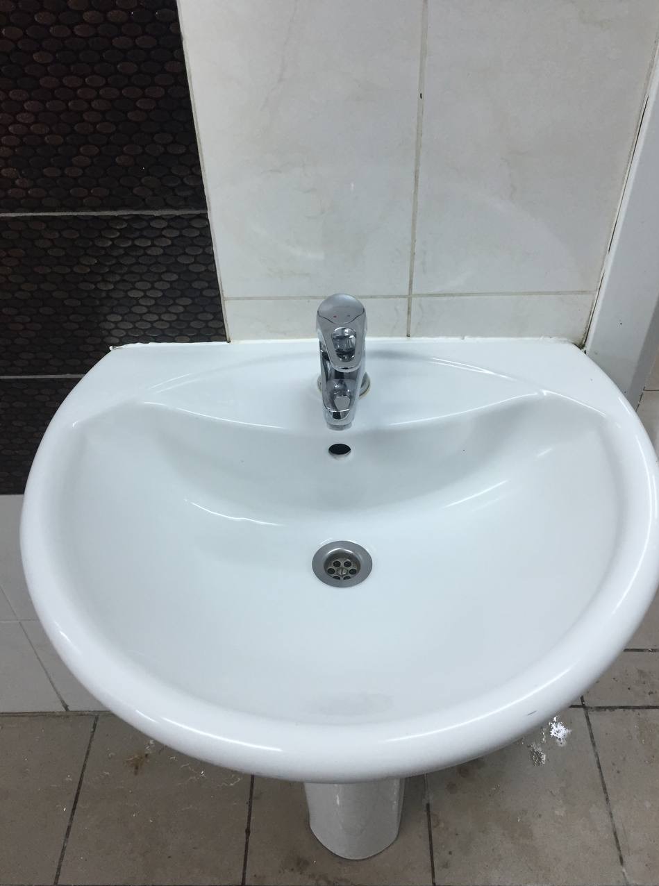 Biała umywalka z chromowaną baterią na tle płytek. Widoczne zabrudzenia na fugach i podłodze, co sugeruje potrzebę sprzątania.