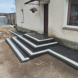 Zuchon usługi ogólnobudowlane - Nowe, czarno-białe schody wejściowe z kostki brukowej prowadzące do domu z wiszącą donicą. Widok na elewację i fragment podwórka z kostką.