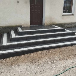 Zuchon usługi ogólnobudowlane - Wykończone schody wejściowe do domu, wyłożone ciemną kostką z jasnymi akcentami. Widoczne kwiaty w doniczce nad drzwiami i fragment elewacji. Estetyczne i nowoczesne.