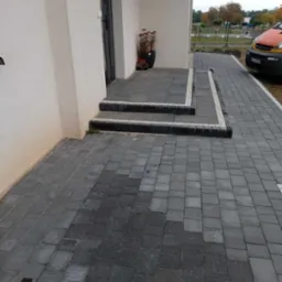 Ułożona kostka brukowa w odcieniach szarości prowadząca do wejścia domu z donicą. Widoczne schody z kostki z białymi obrzeżami i fragment auta.