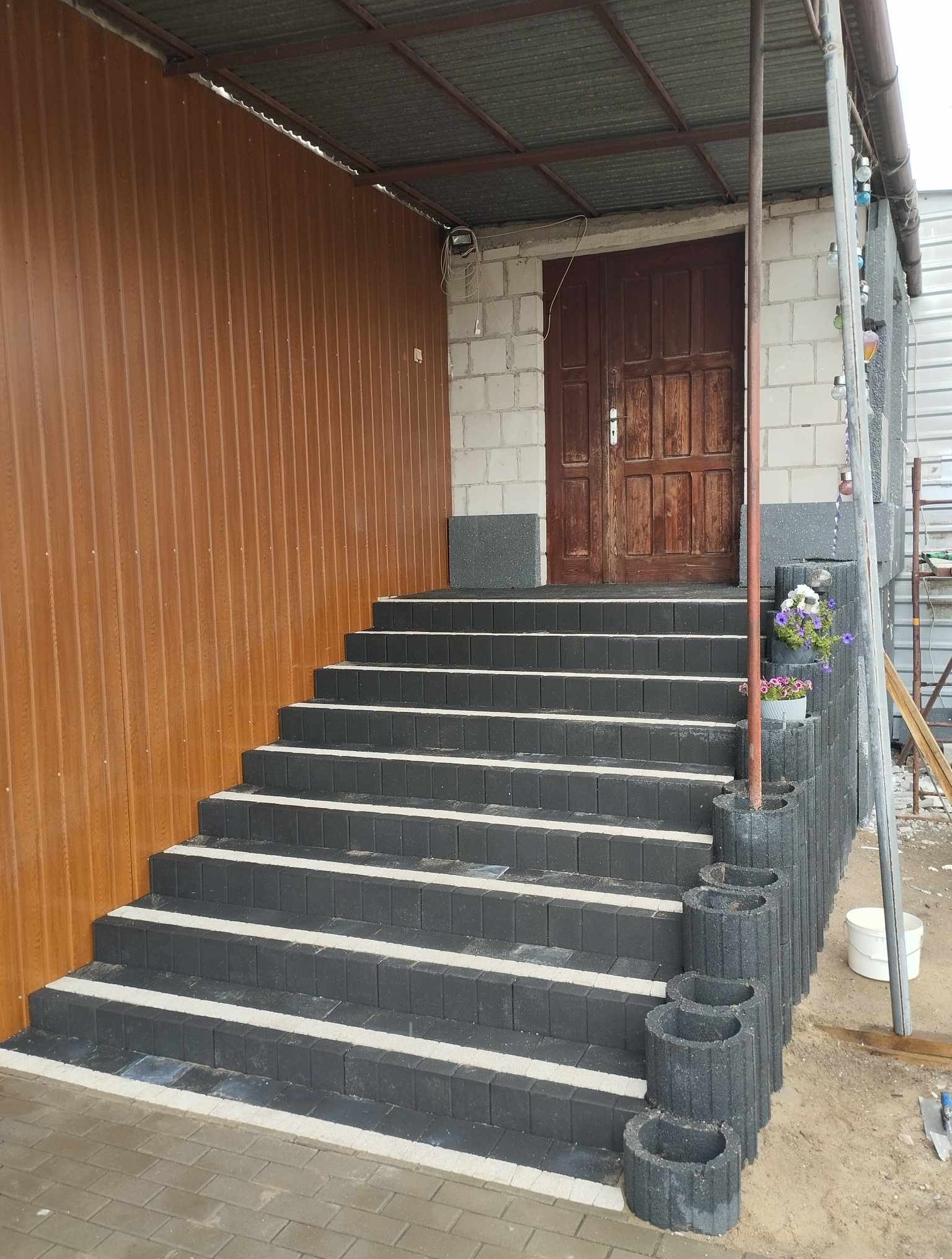 Schody wejściowe z ciemnej kostki z białymi pasami, prowadzące do drewnianych drzwi. Ściana z brązowego panelu i surowego muru. Donica z kwiatami przy schodach.