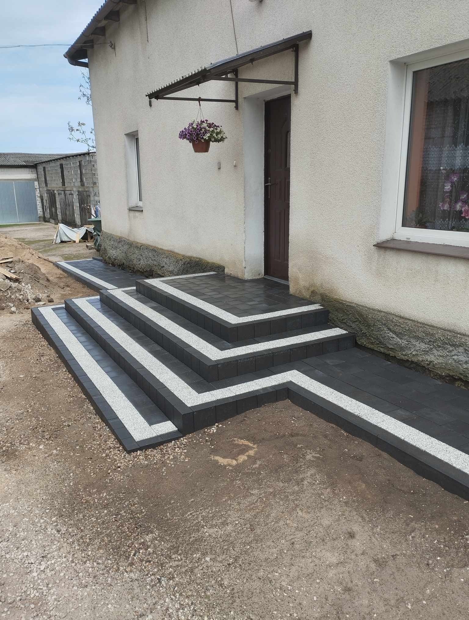 Nowe, czarno-białe schody wejściowe z kostki brukowej prowadzące do domu z wiszącą donicą. Widok na elewację i fragment podwórka z kostką.