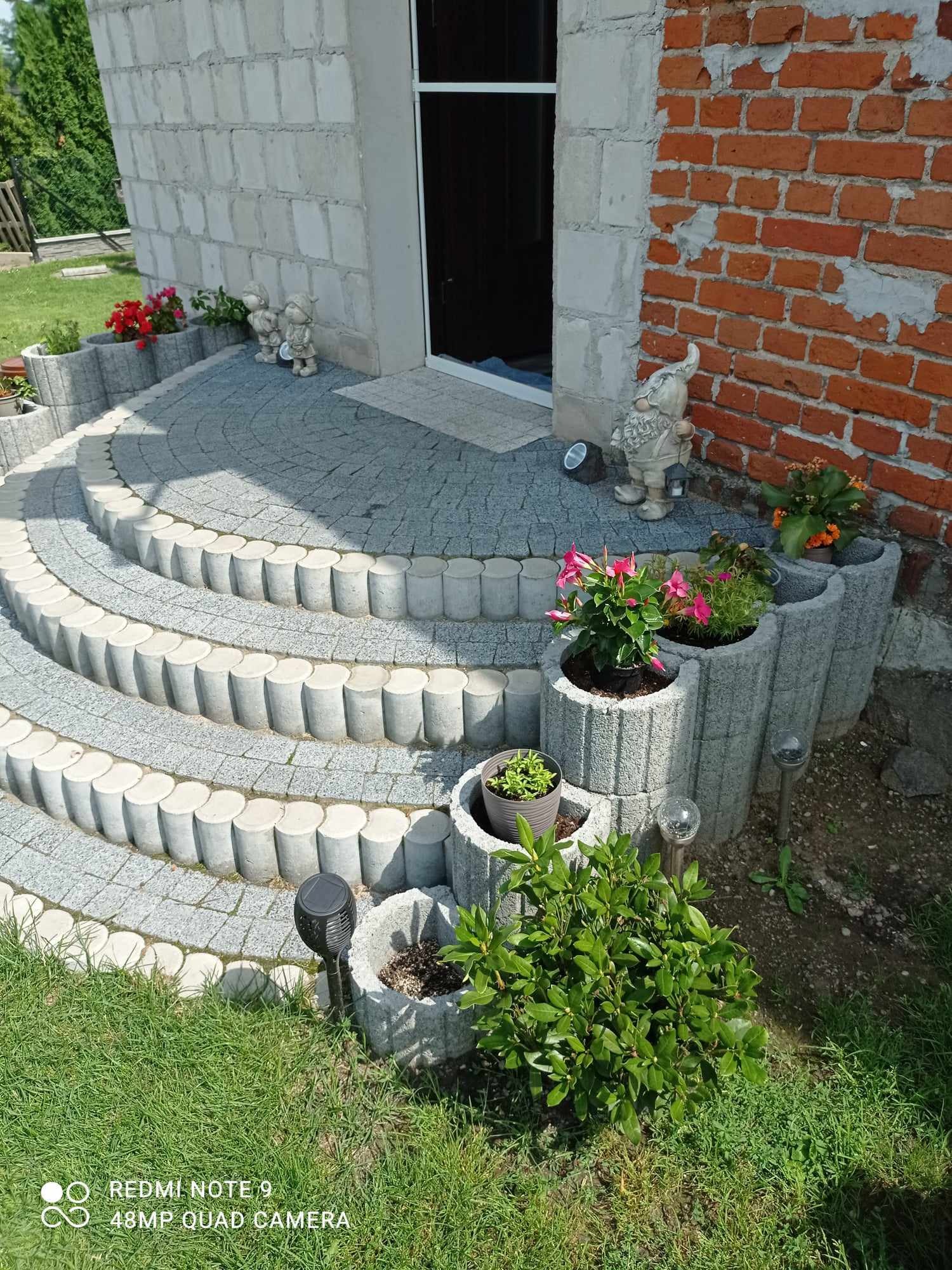 Schody z kostki brukowej prowadzące do wejścia domu, z dekoracyjnymi donicami i figurkami ogrodowymi, na tle ceglanej ściany.