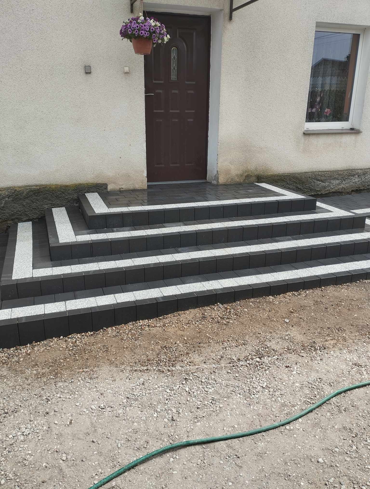 Wykończone schody wejściowe do domu, wyłożone ciemną kostką z jasnymi akcentami. Widoczne kwiaty w doniczce nad drzwiami i fragment elewacji. Estetyczne i nowoczesne.