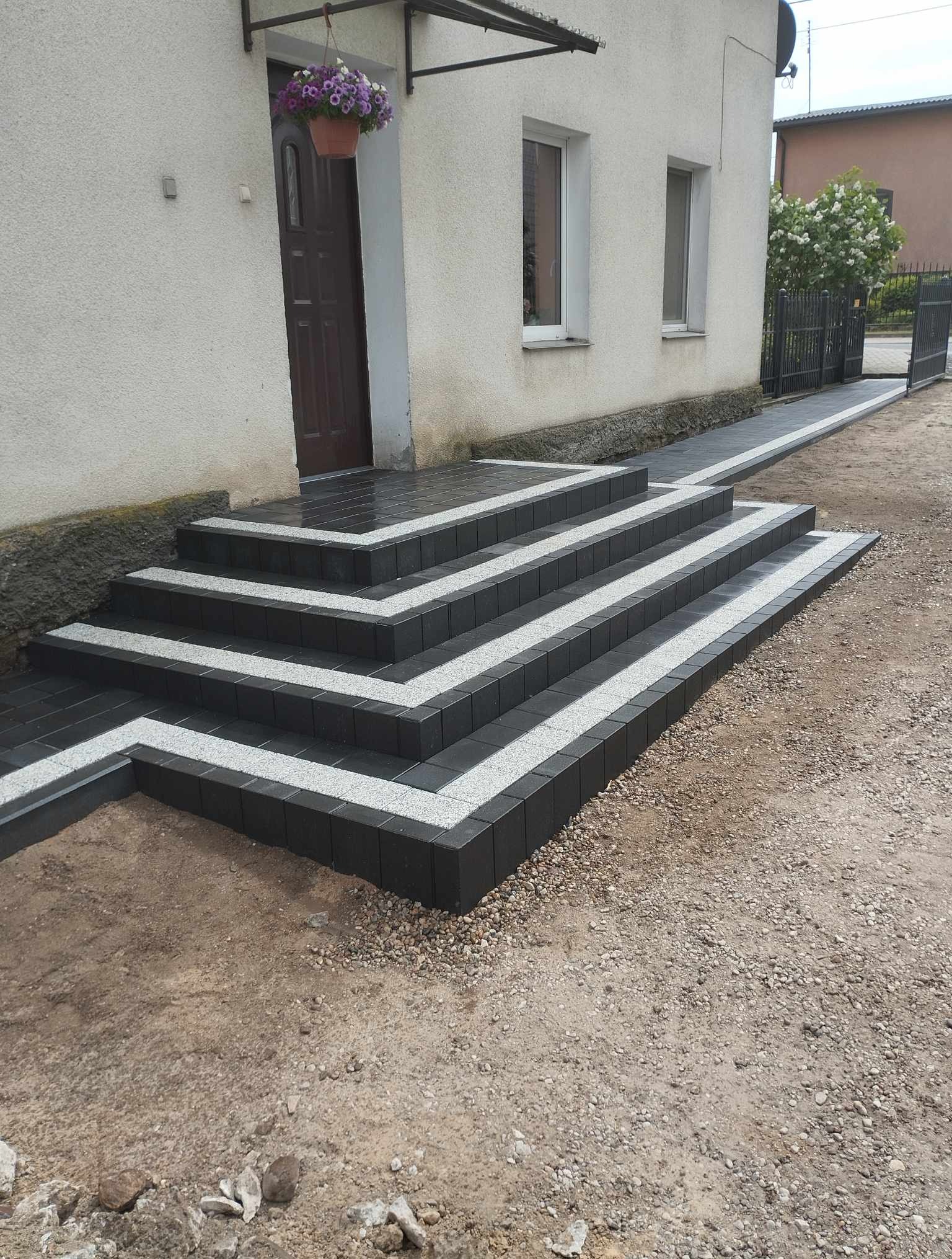 Ułożone schody wejściowe z kostki brukowej w odcieniach szarości, prowadzące do domu z kwiatami w doniczce nad drzwiami. Widoczny chodnik z kostki i teren wokół w trakcie zagospodarowania.