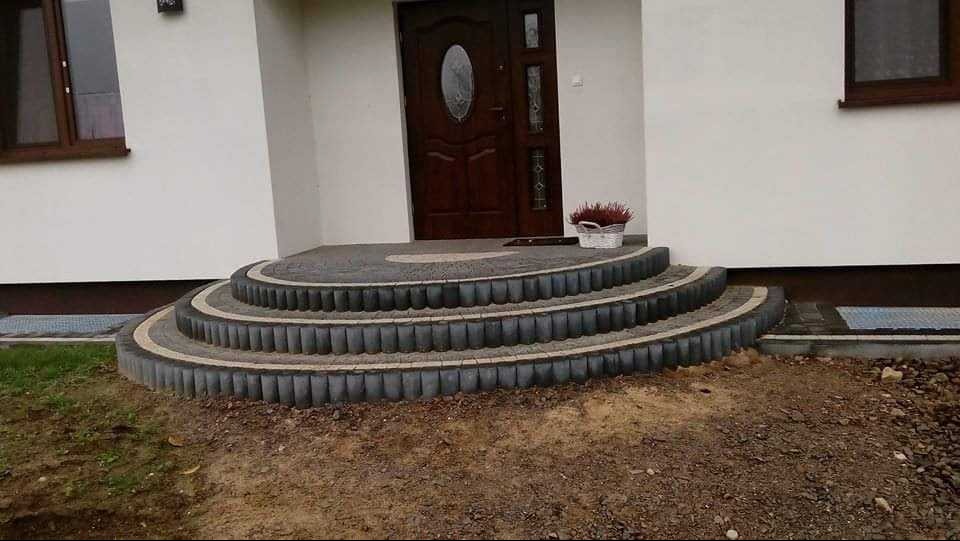 Nowe, trzystopniowe schody wejściowe z kostki brukowej prowadzące do domu z brązowymi drzwiami. Na górnym stopniu stoi koszyk z wrzosem.