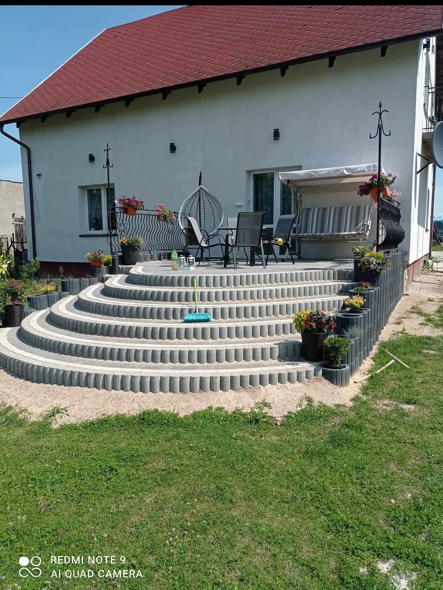 Schody tarasowe z betonowych elementów prowadzące do domu z czerwonym dachem. Widoczne kwiaty w donicach i meble ogrodowe na tarasie. Elementy dekoracyjne.