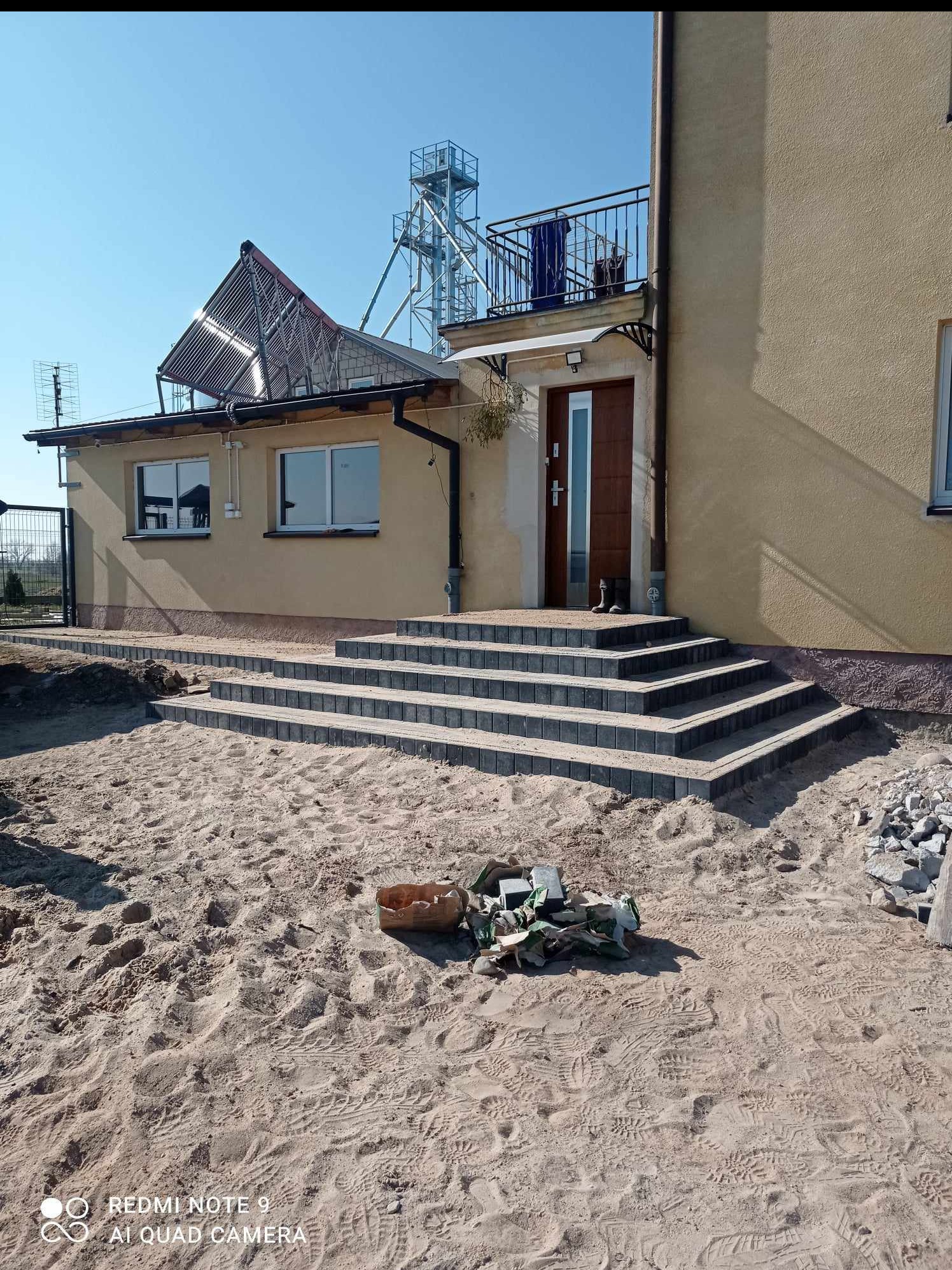 Ułożone schody z kostki brukowej prowadzące do domu z balkonem i panelami słonecznymi na dachu. Na pierwszym planie piasek i resztki materiałów budowlanych.