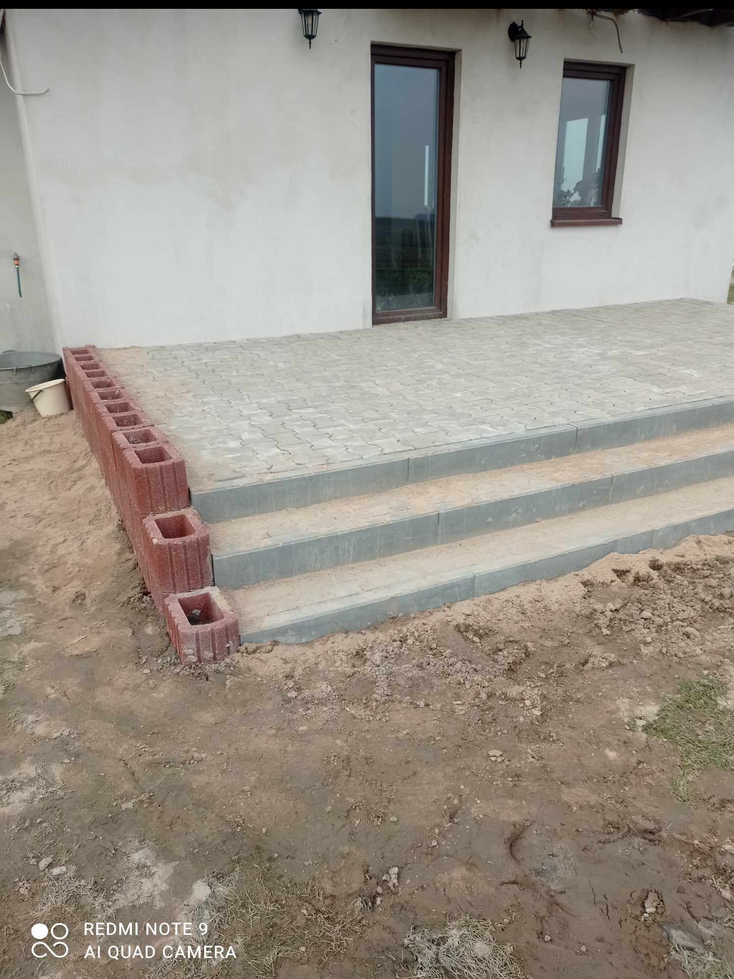 Wykończone schody wejściowe do domu z kostki brukowej i obrzeżem z czerwonych pustaków. Widoczne drzwi i okno domu w tle. Prace ziemne w toku.
