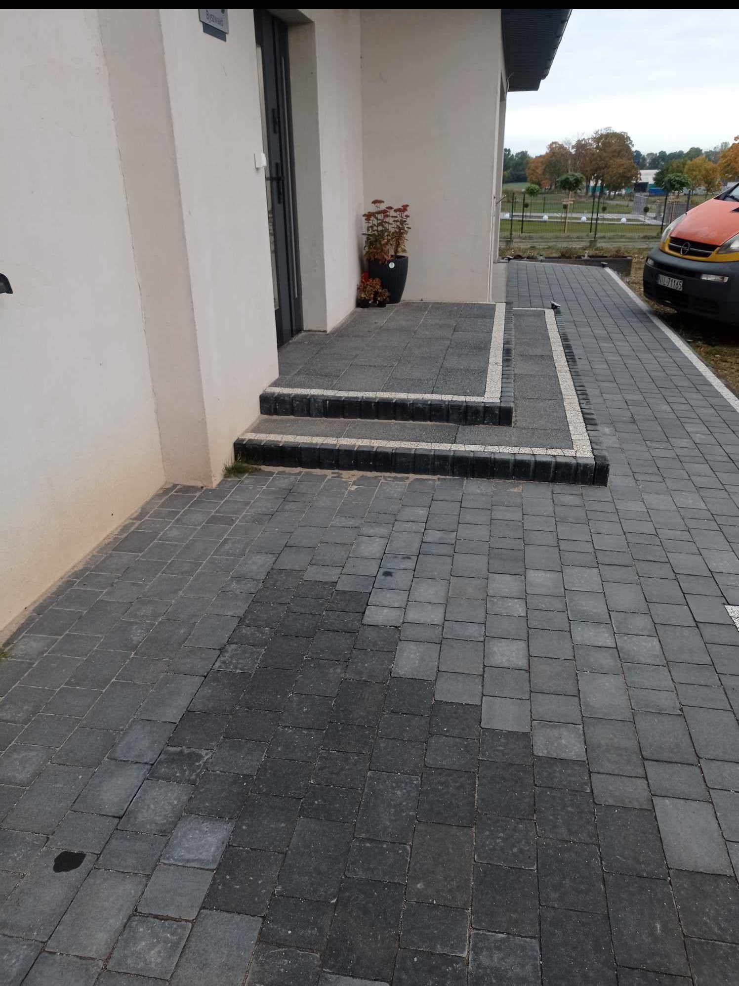 Ułożona kostka brukowa w odcieniach szarości prowadząca do wejścia domu z donicą. Widoczne schody z kostki z białymi obrzeżami i fragment auta.