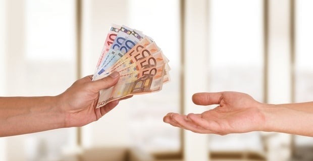 Dłoń wręcza plik banknotów euro drugiej dłoni na rozmytym tle. Nominały widoczne: 10, 20, 50. Symboliczne przekazanie środków finansowych.