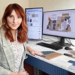 Krakowska Projektownia - Kobieta z rudymi włosami prezentuje próbki materiałów na tle monitorów z projektami wnętrz. Widoczny plan mieszkania i moodboard.