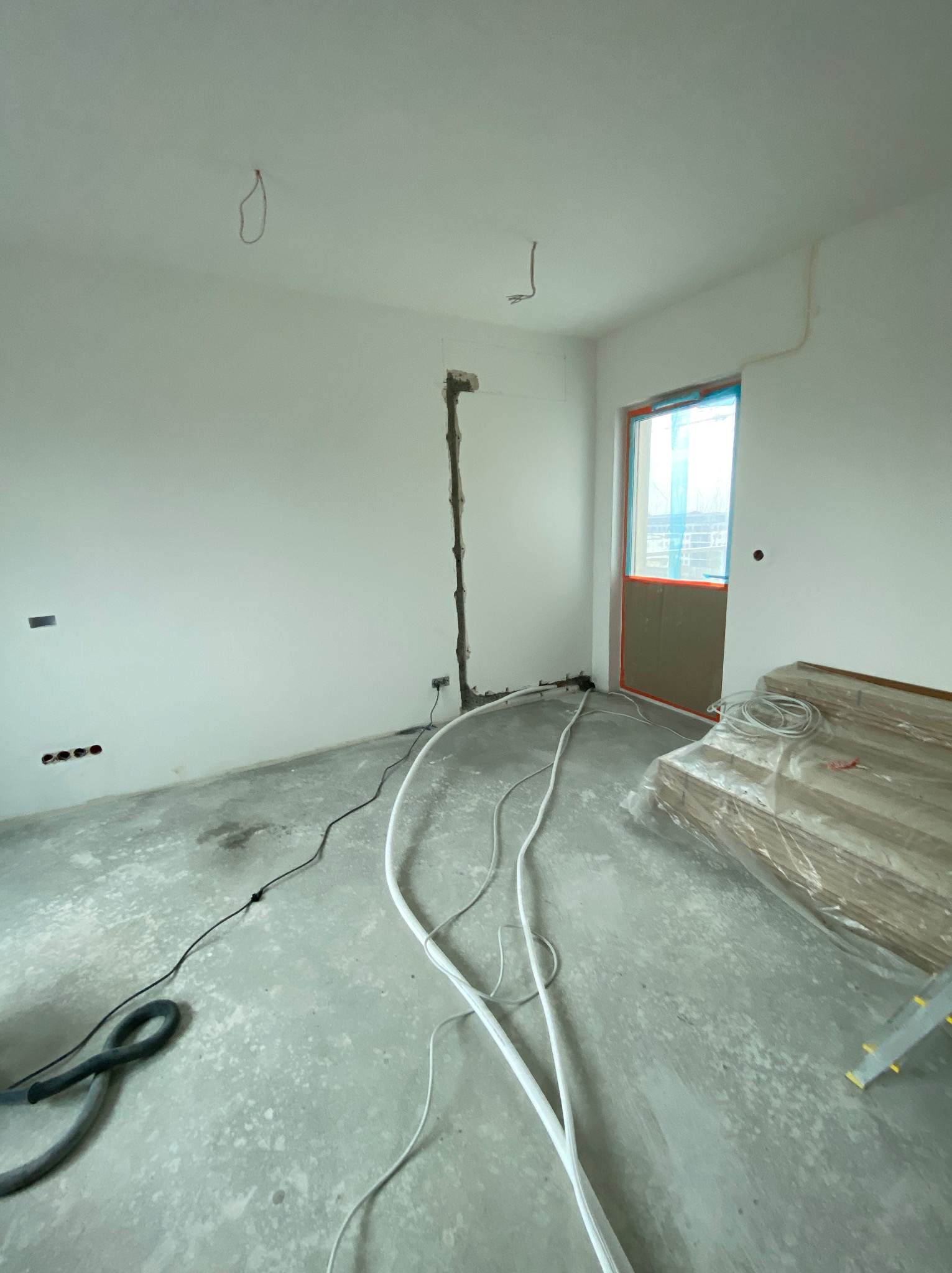 Surowy stan wnętrza z przewodami i rurami na podłodze, przygotowanego do instalacji grzewczej i klimatyzacji. Widoczne puszki elektryczne i drzwi z zabezpieczeniem.