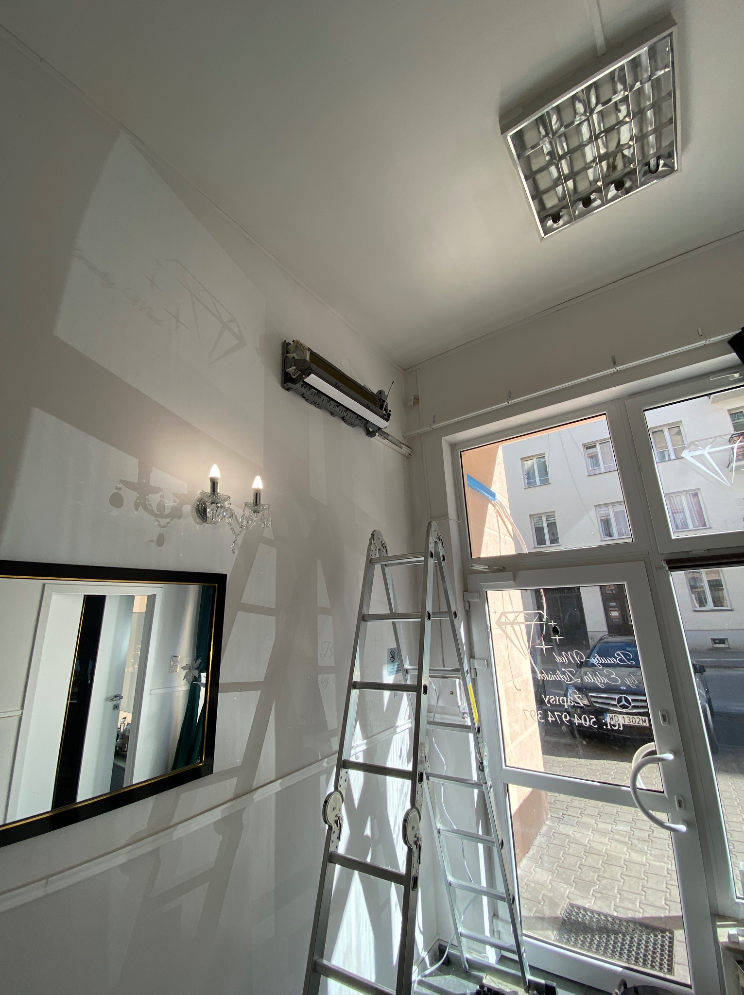 Wnętrze z klimatyzatorem na ścianie, drabina oparta o ścianę, widok przez okno na budynek i samochód. Oświetlenie sufitowe i kinkiet.