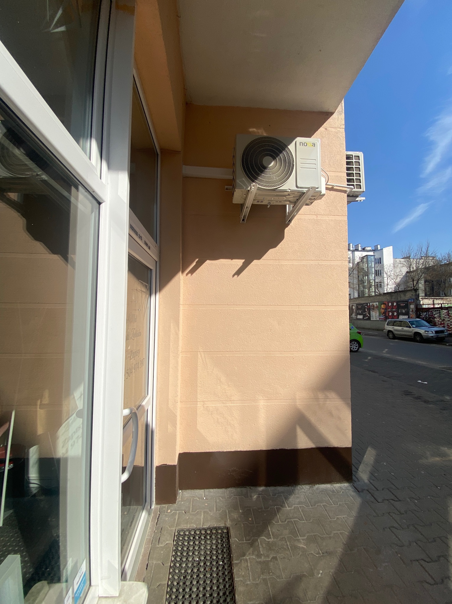 Zamontowana klimatyzacja Nova na elewacji budynku z widokiem na ulicę i błękitne niebo. Widoczny fragment okna i drzwi wejściowych.