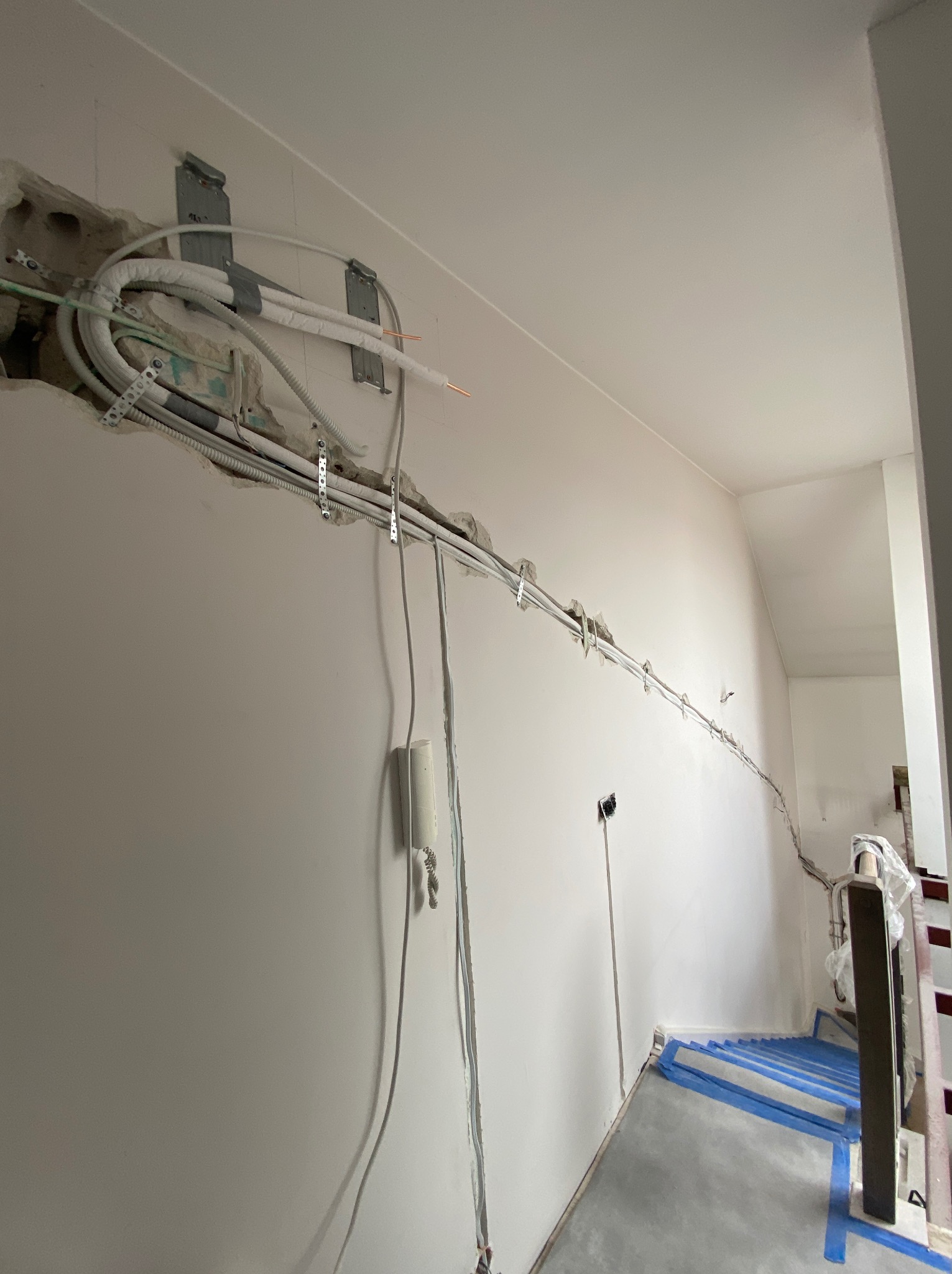Instalacja przewodów klimatyzacji w bruzdach ściennych, widoczne kable i rury zabezpieczone taśmą, fragment schodów z zabezpieczeniem. Prace w trakcie realizacji.