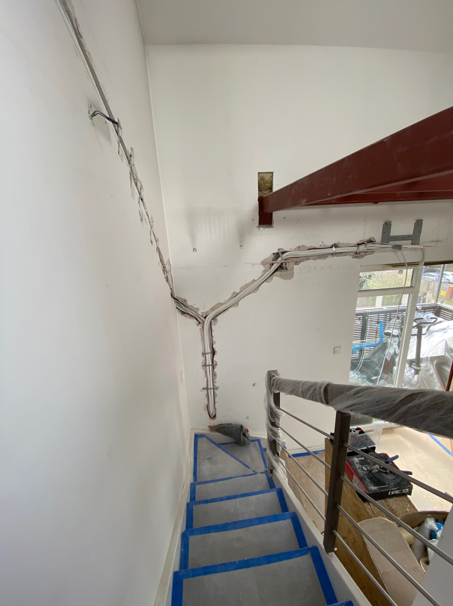 Instalacja elektryczna w bruzdach na ścianie, widoczne przewody i kable. Schody zabezpieczone folią malarską, częściowo widoczna stalowa belka stropowa.