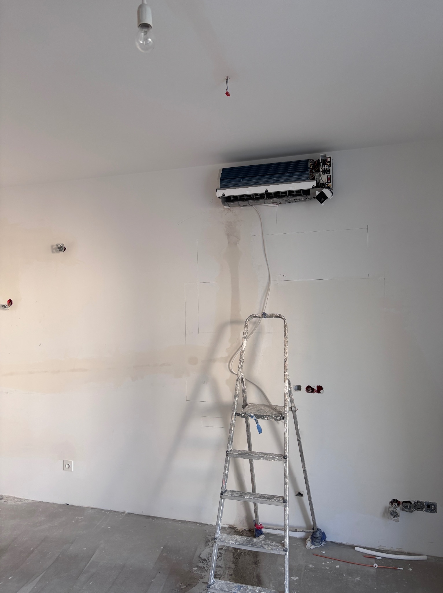 Montaż klimatyzacji na ścianie w trakcie remontu. Widoczna drabina, wystające kable i niedokończone wykończenie ściany. Klimatyzator w trakcie instalacji.