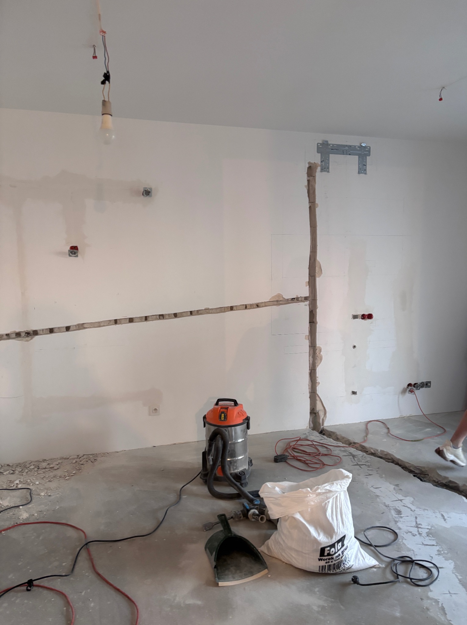 Przygotowanie pod instalację klimatyzacji: bruzdy w ścianie, wspornik montażowy, odkurzacz przemysłowy i worek gruzu na szarym, betonowym podłożu.
