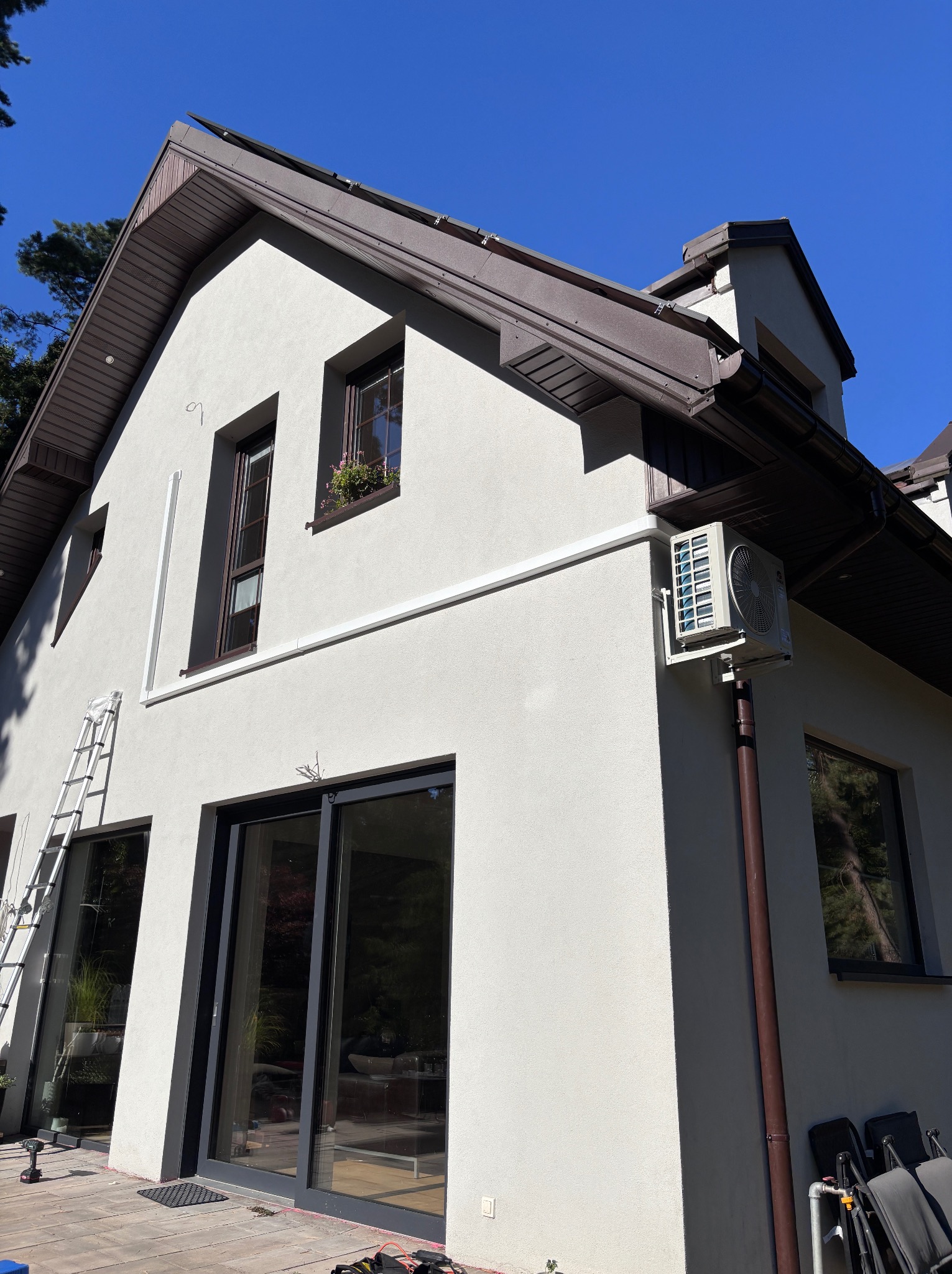 Elewacja domu z zamontowaną jednostką zewnętrzną klimatyzacji. Widoczna drabina i narzędzia sugerują trwający montaż. Jasna elewacja i brązowe detale.