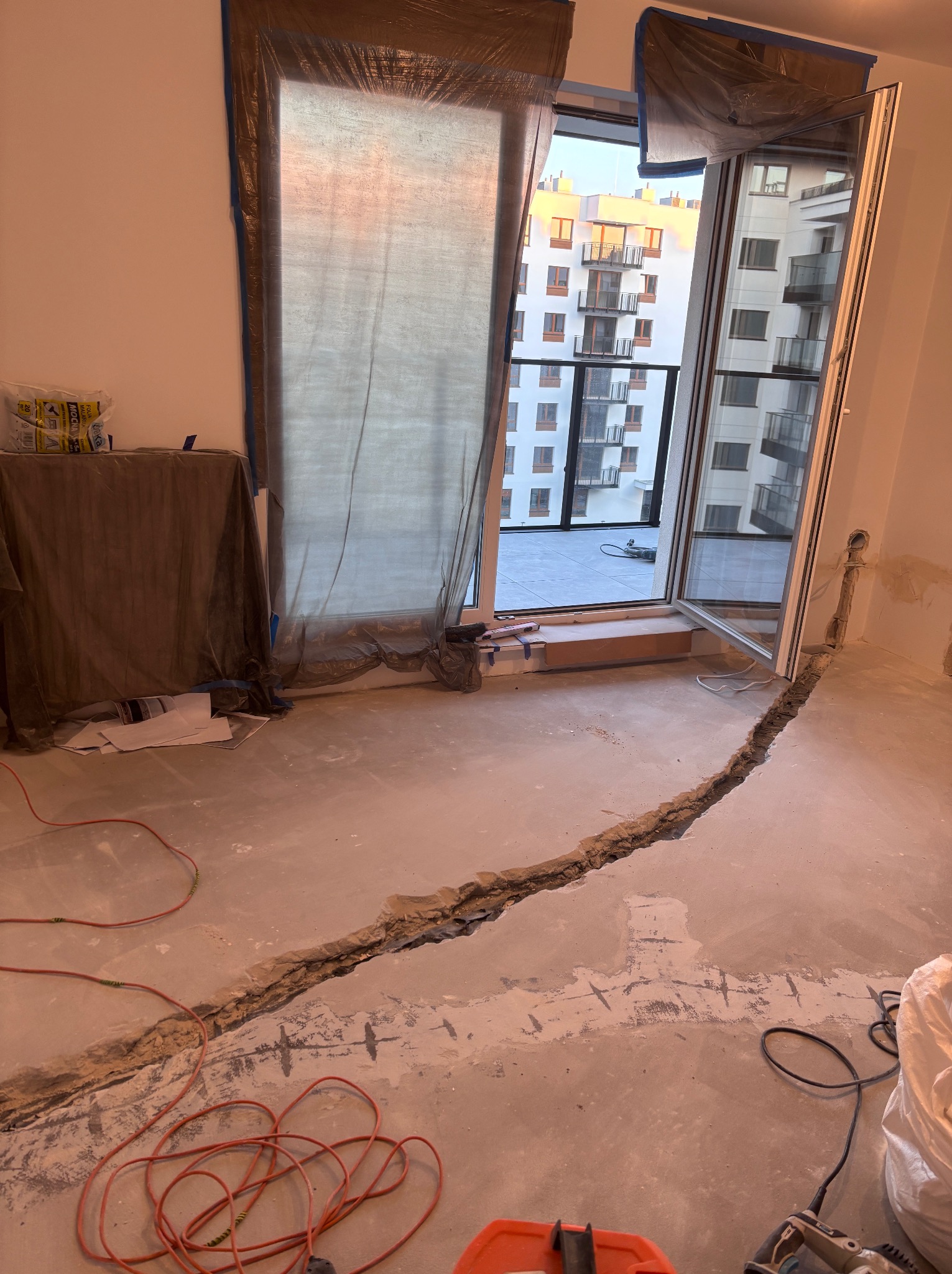 Przygotowanie pod instalację grzewczą w pomieszczeniu: wycięty rowek w podłodze, zabezpieczone okno, widoczne narzędzia i kable. Etap prac instalacyjnych.