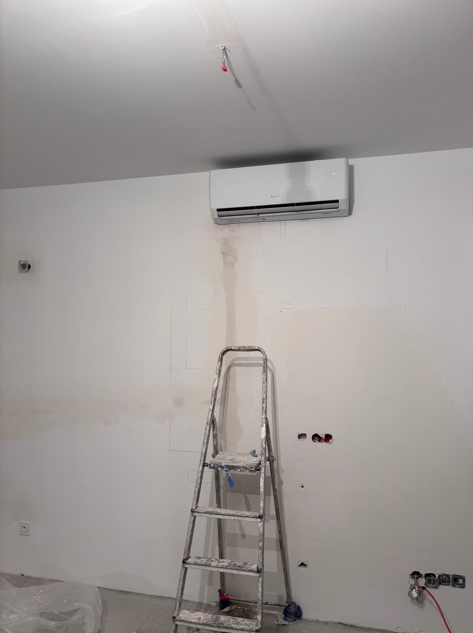 Biała klimatyzacja na ścianie w trakcie remontu, widoczna drabina malarska i ślady po pracach wykończeniowych. Oświetlenie sufitowe, surowy stan ścian.